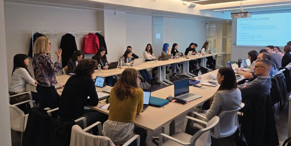 un gruppo di persone giovani sono sedute in una sala riunioni e ascoltano una docente che parla
