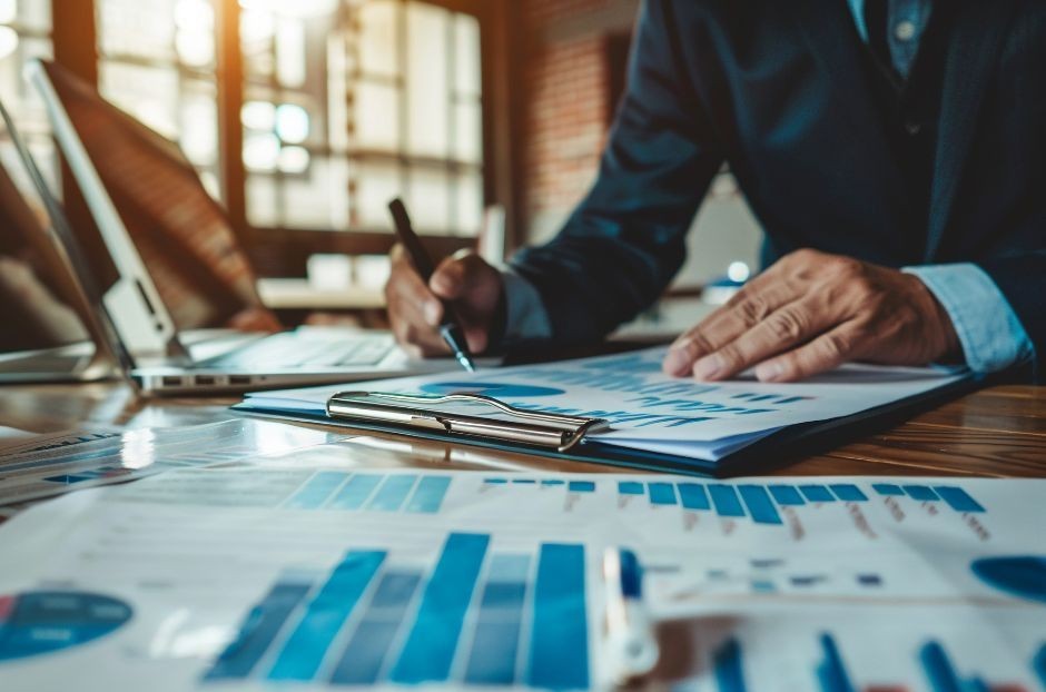 un uomo, seduto ad un tavolo con una penna in mano, controlla un documento con grafici di performance